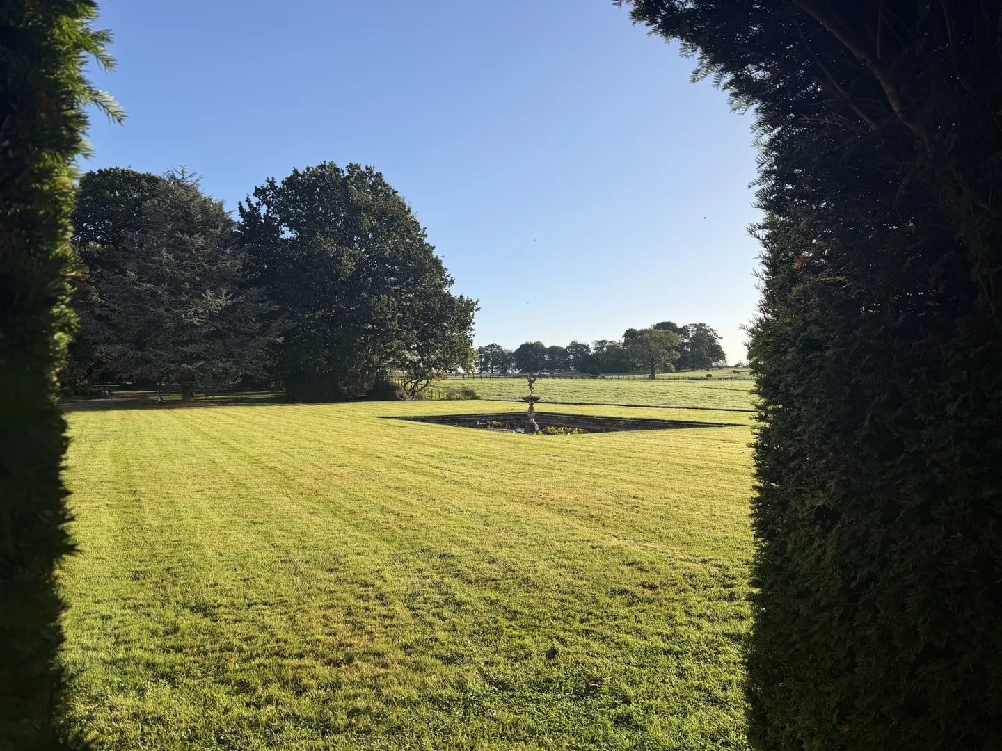 Striped lawn and garden fountain framed by yew hedges, with parkland beyond